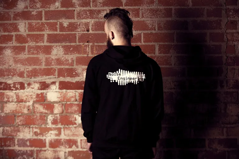 An extremely good-looking person facing a brick wall wearing a hoodie with the Noise Engineering logo on the back.